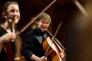 Juge Musiker:innen in dunkler Kleidung mit Streichinstrumenten, die in die Kamera lächeln.