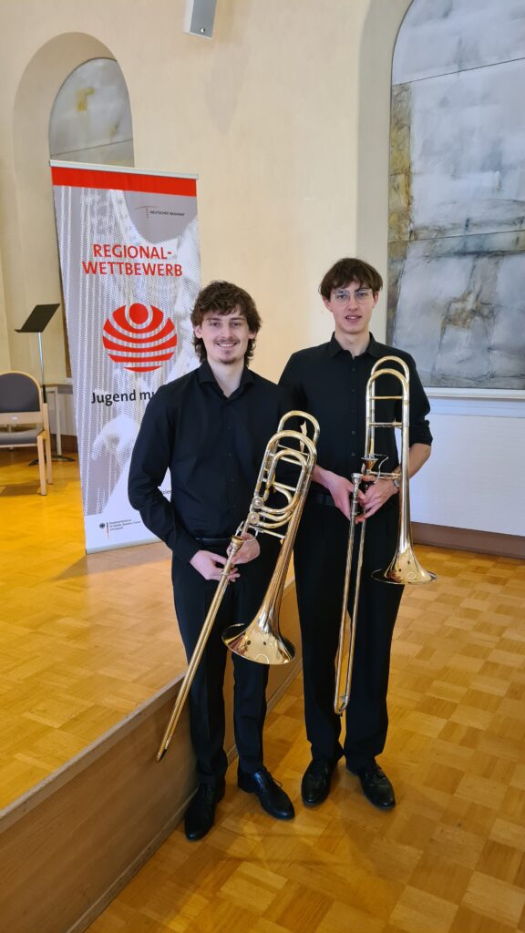 Preisträger Marius Munz und Benedikt Kasper mit Posaunen vor einer Bühne in einem Konzertsaal.