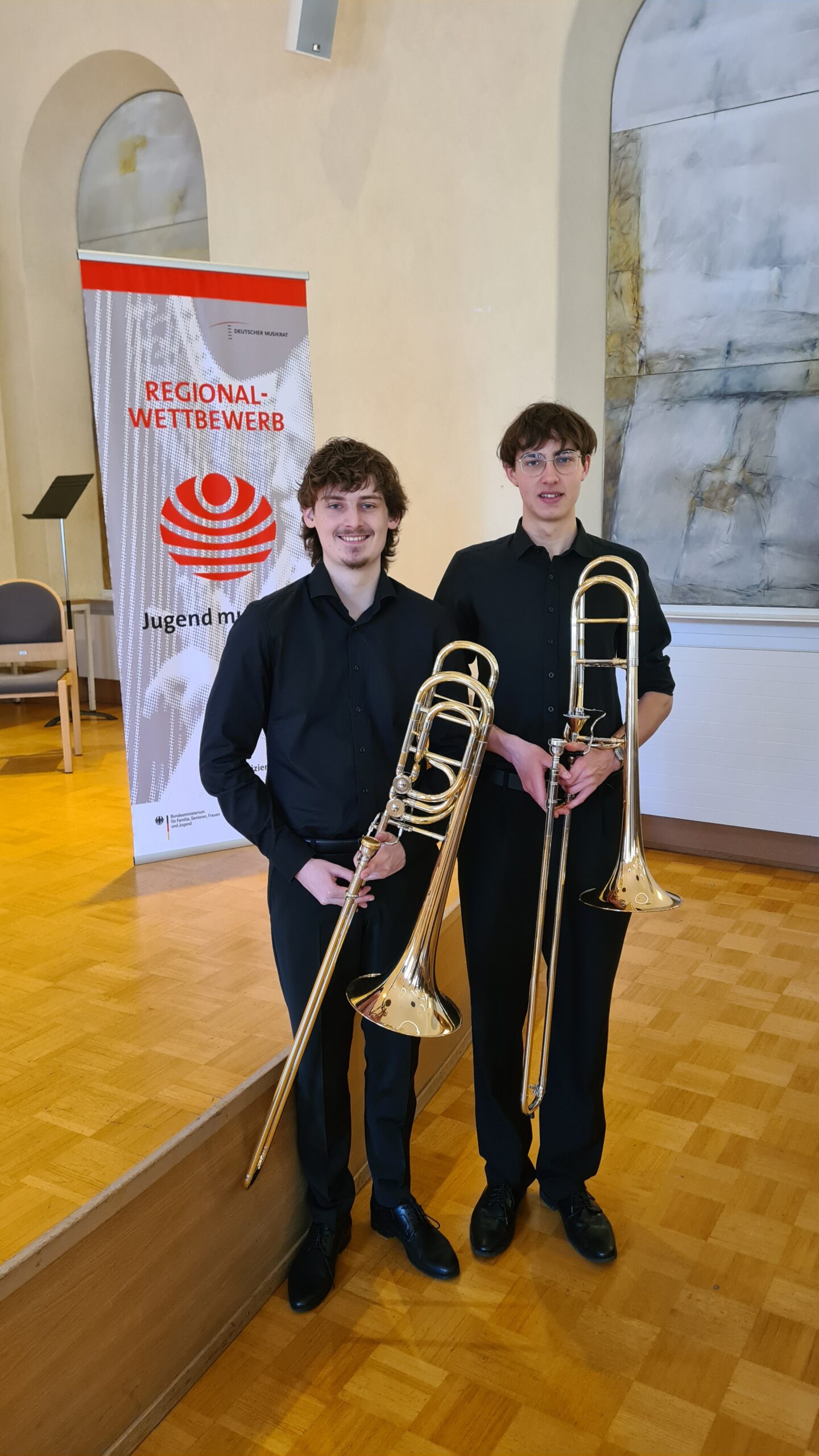 Preisträger Marius Munz und Benedikt Kasper mit Posaunen vor einer Bühne in einem Konzertsaal.