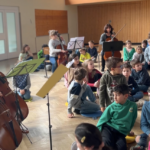 Kinder und Lehrerin spielen im Streicherensemble. Dazwischen sitzen Kinder, die zuhören